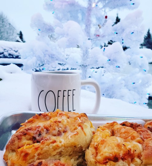A cozy winter scene featuring a mug of coffee and freshly baked cinnamon rolls in front of a snowy white tree.