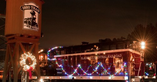 A decorated train at night, featuring festive lights and a wreath, with a water tower in the background.