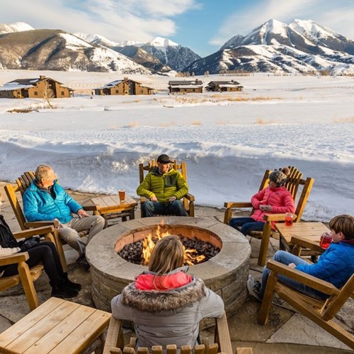 A group of people sitting around a fire pit in a snowy landscape, with mountains and cabins in the background.