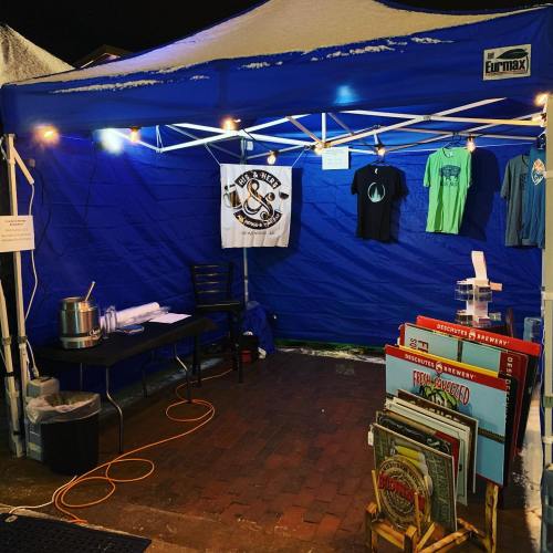 A blue tent with shirts hanging, a table, and a display of beer signs, illuminated by string lights at night.