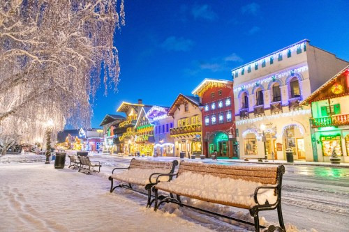 Snow-covered street in a festive town, lined with colorful, illuminated buildings and benches under a blue sky.