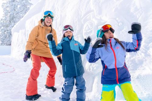 A woman and two children joyfully pose in the snow, wearing colorful winter gear, with a snow sculpture in the background.