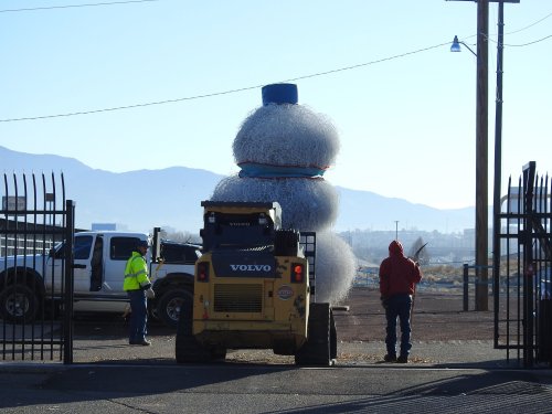 A construction vehicle lifts a large, round object while two workers observe in an outdoor setting. Mountains in the background.