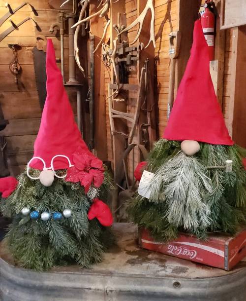 Two festive gnome decorations made of greenery, wearing red hats, glasses, and adorned with holiday accents.