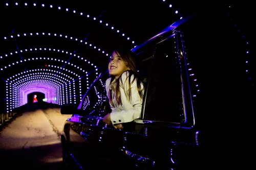 A girl leans out of a car window, smiling in a colorful, illuminated tunnel.