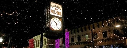 A clock tower in a festive village, surrounded by colorful holiday lights and falling snow at night.