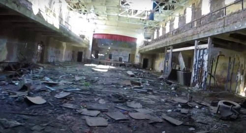 An abandoned, dilapidated interior of a large hall with debris scattered across the floor and sunlight streaming in.