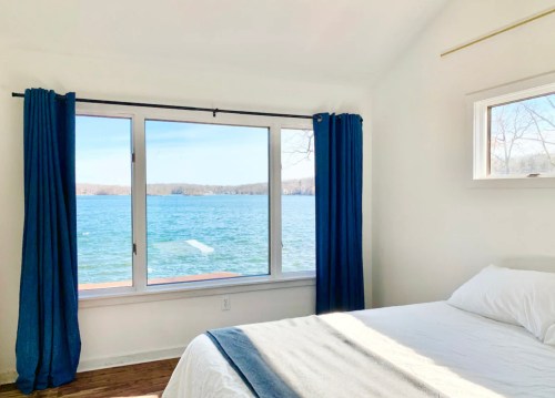 Bright bedroom with a large window overlooking a serene lake, featuring white walls and blue curtains.