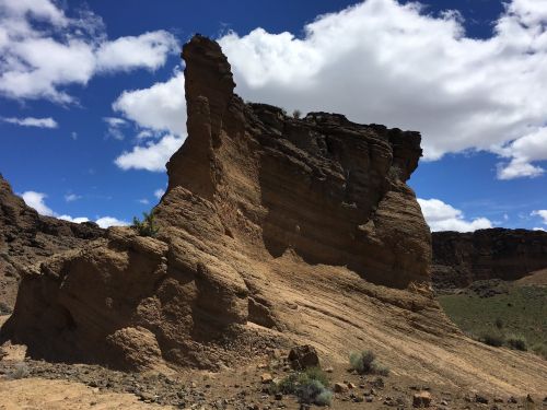 A tall, rugged rock formation stands against a blue sky with fluffy clouds, surrounded by arid landscape.