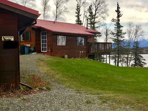 A wooden cabin with a red roof, surrounded by trees and grass, near a lake under a cloudy sky.
