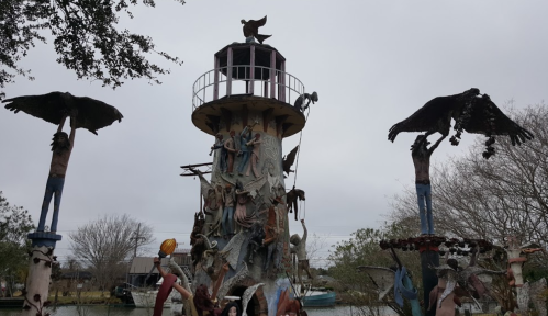 A whimsical lighthouse surrounded by sculptures of birds and figures, set against a cloudy sky.