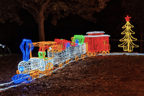A colorful light display of a train carrying gifts next to a decorated Christmas tree.