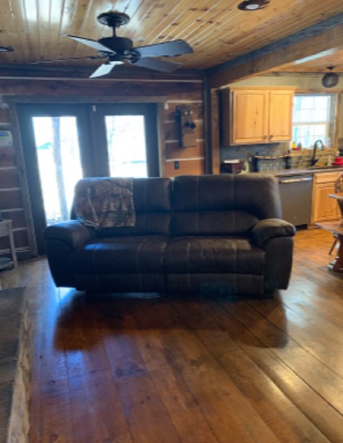 Cozy living room with a brown couch, wooden floors, and a rustic kitchen in the background. Natural light from the windows.