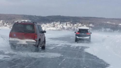 Two vehicles driving on a snowy road with strong winds and snow drifts, surrounded by a winter landscape.