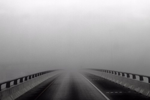 A foggy road stretches into the distance, obscured by thick mist, with guardrails on either side.
