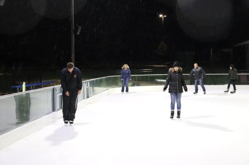 People ice skating on an outdoor rink at night, with snow falling and lights illuminating the scene.