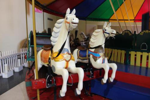 Two vintage carousel horses with colorful saddles, set against a bright, multicolored tent.