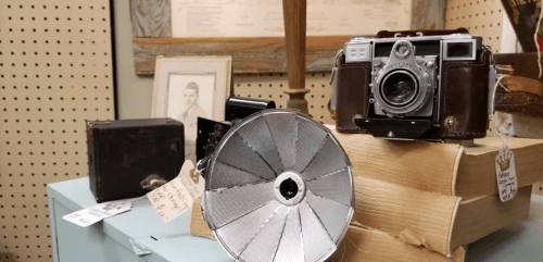 A vintage camera and flash sit atop stacked books in an antique shop, with framed art in the background.