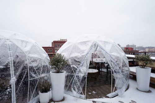 Two transparent igloos covered in snow on a rooftop, with potted plants nearby and a cityscape in the background.