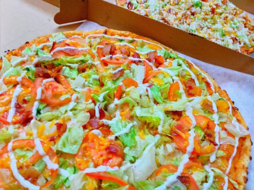 A close-up of a fresh pizza topped with lettuce, tomatoes, cheese, and drizzled with mayonnaise, in a pizza box.
