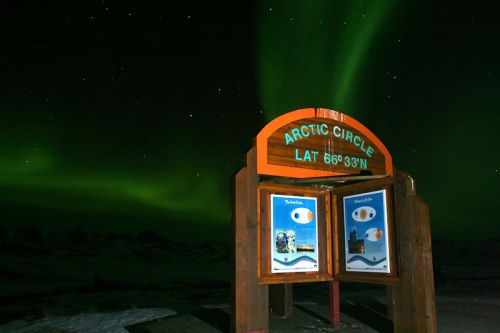 Sign marking the Arctic Circle under a vibrant display of the Northern Lights in a starry night sky.