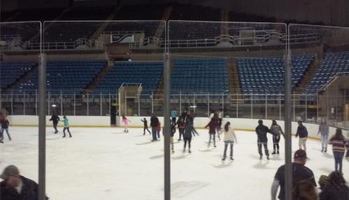 A crowded ice rink with people skating, surrounded by empty blue seats in a sports arena.