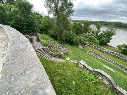 A stone pathway leads down to a riverbank, surrounded by lush greenery and cloudy skies.