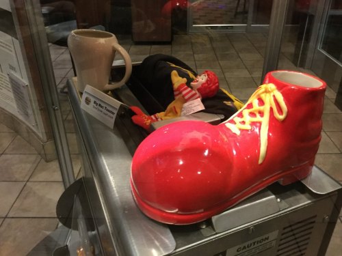 A large red clown shoe and a mug sit on a display, alongside a small Ronald McDonald plush toy.