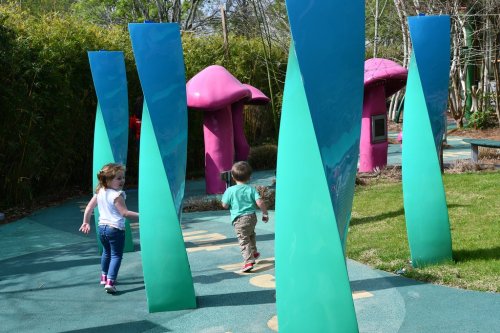 Two children play in a colorful park, walking between tall, reflective green structures and vibrant mushroom-shaped play areas.