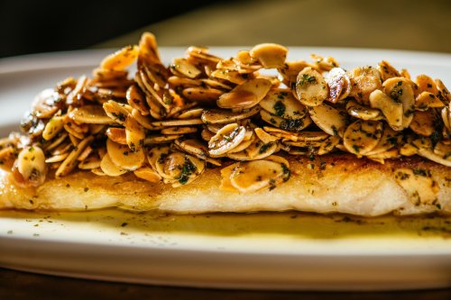 A beautifully plated fish fillet topped with toasted almonds and herbs, drizzled with olive oil on a white dish.