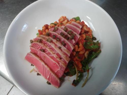 Sliced seared tuna on a bed of greens and shrimp, garnished with capers in a white bowl.