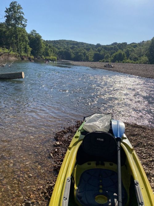 A kayak on a rocky riverbank with clear water and lush green hills in the background under a bright blue sky.