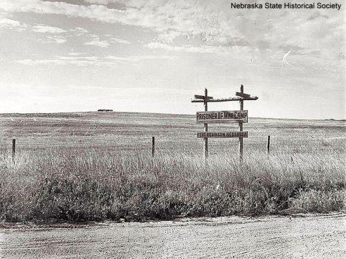 Sign for a prisoner war camp in Nebraska, set against a vast, open landscape with a cloudy sky.