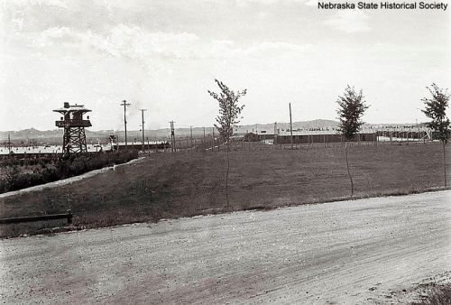 Black and white historical photo of a watchtower overlooking a fenced area, with a dirt road in the foreground.