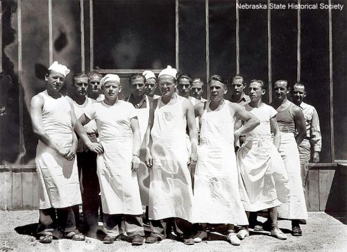 A group of male bakers in white aprons and hats, posing together outside a building.