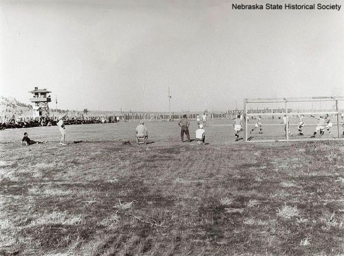 A historical black-and-white photo of a baseball game with spectators in the background and a watchtower nearby.