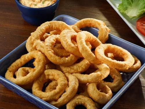 A blue bowl filled with golden, crispy onion rings on a wooden table, with a small bowl of dip in the background.