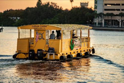 A yellow party boat named "Latitude Lindsey" cruising on a calm waterway in Tampa Bay, adorned with lights.