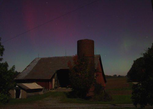 A red barn under a starry sky with auroras, surrounded by trees and fields at night.
