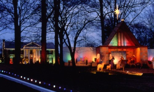 A festive scene featuring a decorated house and a nativity display, illuminated by colorful lights at dusk.
