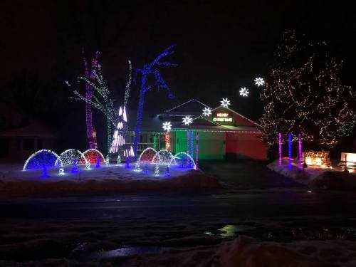 Colorful holiday lights decorate trees and a house, creating a festive winter scene at night. Snow covers the ground.
