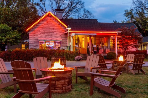 A cozy house with red neon lights, surrounded by fire pits and wooden chairs, set against a twilight sky.