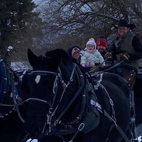 A family smiles while riding in a horse-drawn sleigh through a snowy landscape. A baby is bundled up in the front.
