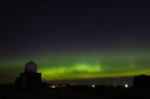 A night sky illuminated by green auroras, with a building silhouette in the foreground and stars twinkling above.