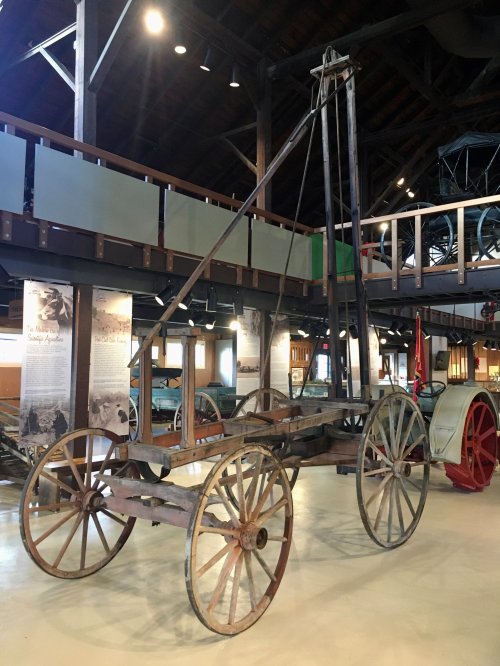 A vintage wooden cart with large wheels, displayed in a museum setting with other historical artifacts in the background.