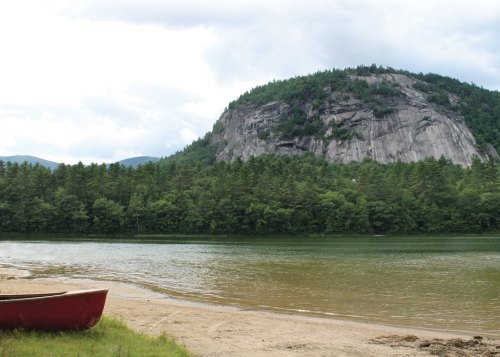 A serene lake with a red canoe on the shore, surrounded by lush trees and a large rocky hill in the background.