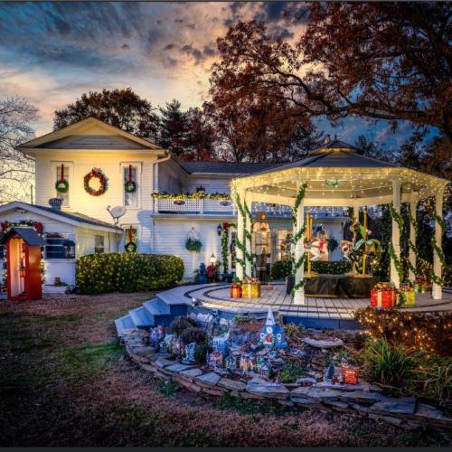A beautifully decorated house with festive lights, a gazebo, and holiday decorations, set against a colorful sunset.