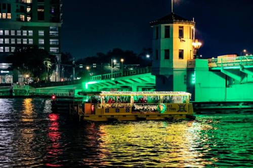 A brightly lit boat glides on the water at night, with a colorful bridge and city lights in the background.
