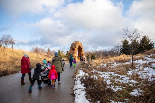 A group of people walks along a path towards a large stone arch, surrounded by grass and snow under a cloudy sky.
