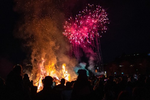 A large bonfire with bright flames, surrounded by a crowd, under a night sky filled with colorful fireworks.